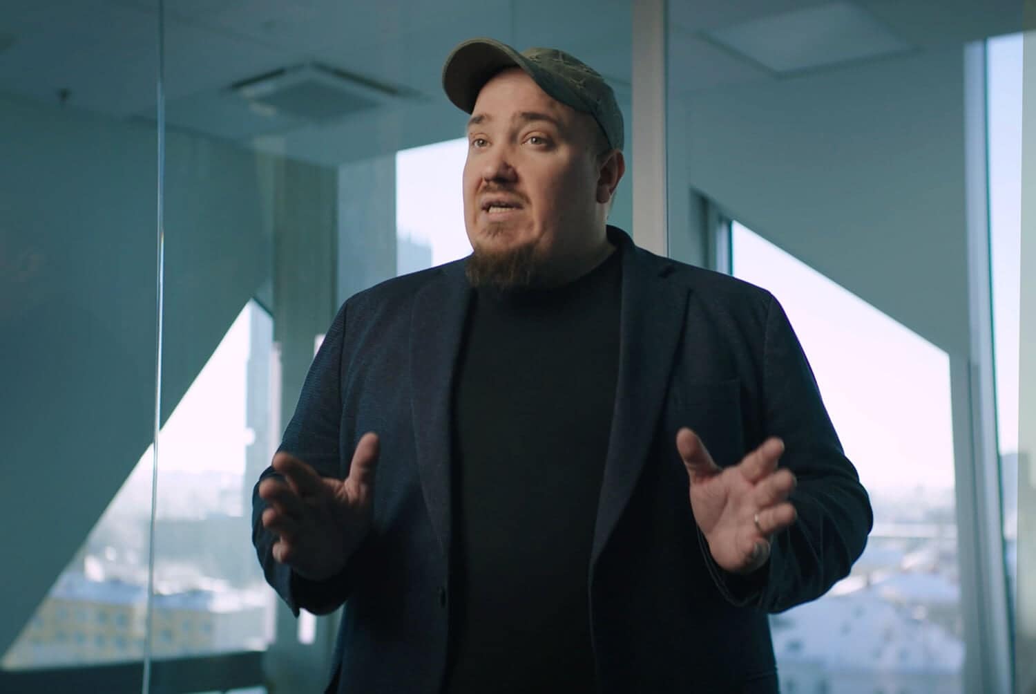 A man with a beard and a baseball cap, wearing a dark blazer and black shirt, gestures with both hands while speaking in a modern office with large windows and city views.