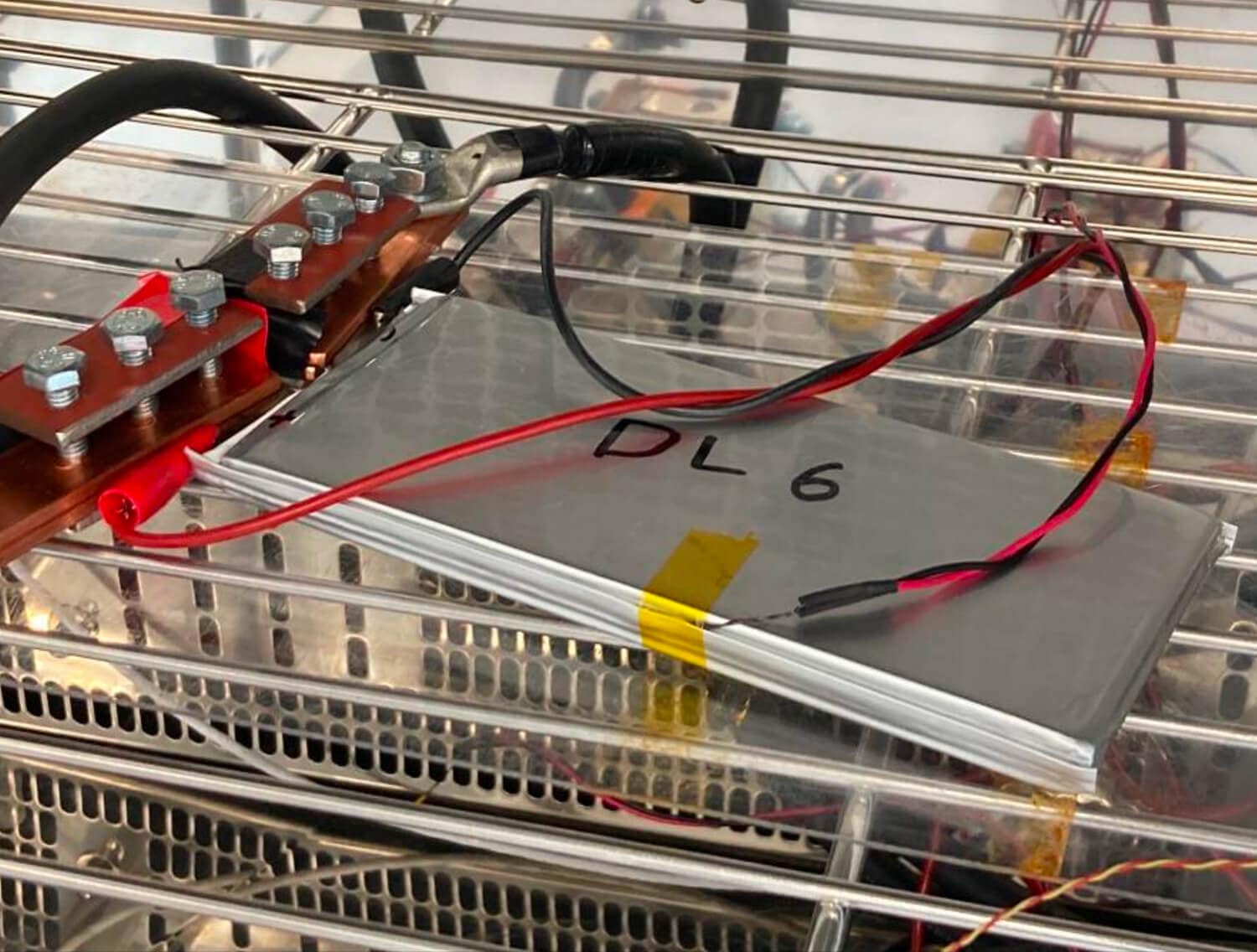 A rectangular battery cell labeled “DL 6” is connected to wires and clips, resting on a metal grate with cables attached to copper terminals above it.