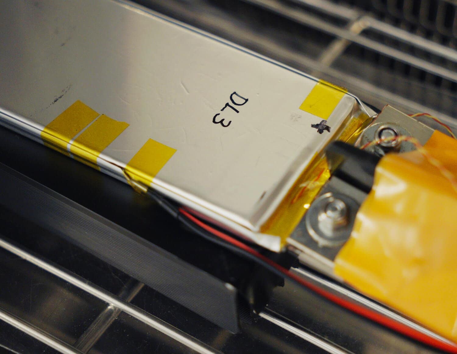 A close-up of a rectangular lithium-ion battery cell with yellow tape on its edges, labeled "D13," connected to wires and metal terminals, resting on a metal rack.