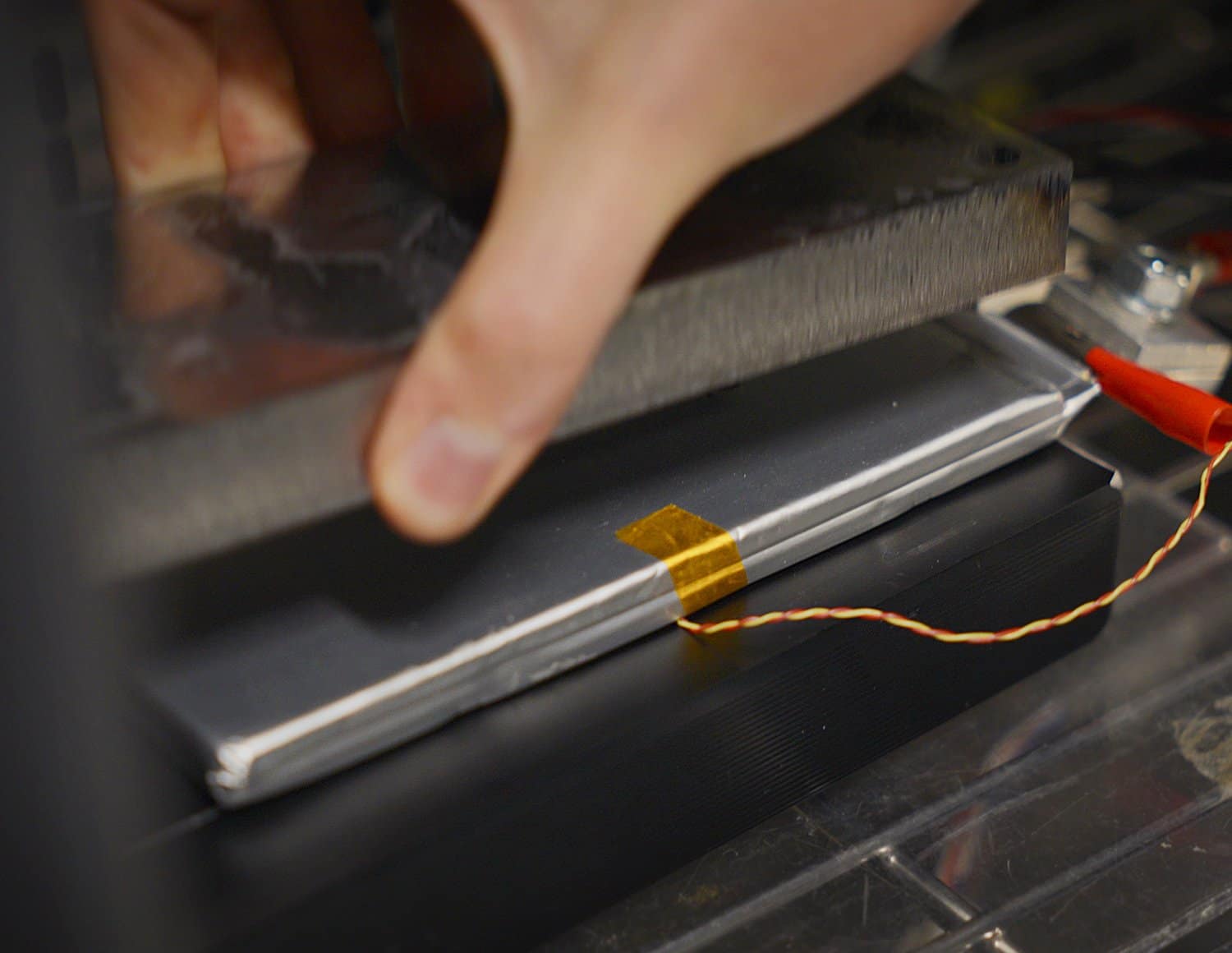 A close-up of a hand pressing a metal plate onto a rectangular battery cell, with a yellow piece of tape and a thin wire attached to the cell, likely for testing or assembly purposes.