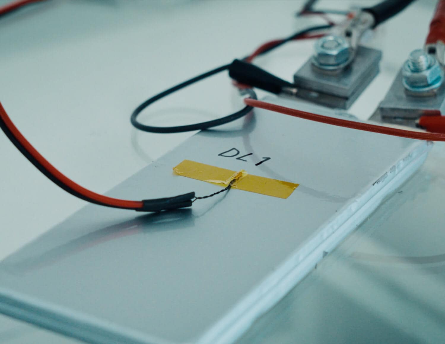 Close-up of an electronic component labeled "DL1" with wires attached using clips and yellow tape, placed on a white surface. The setup appears to be part of an experiment or testing procedure.