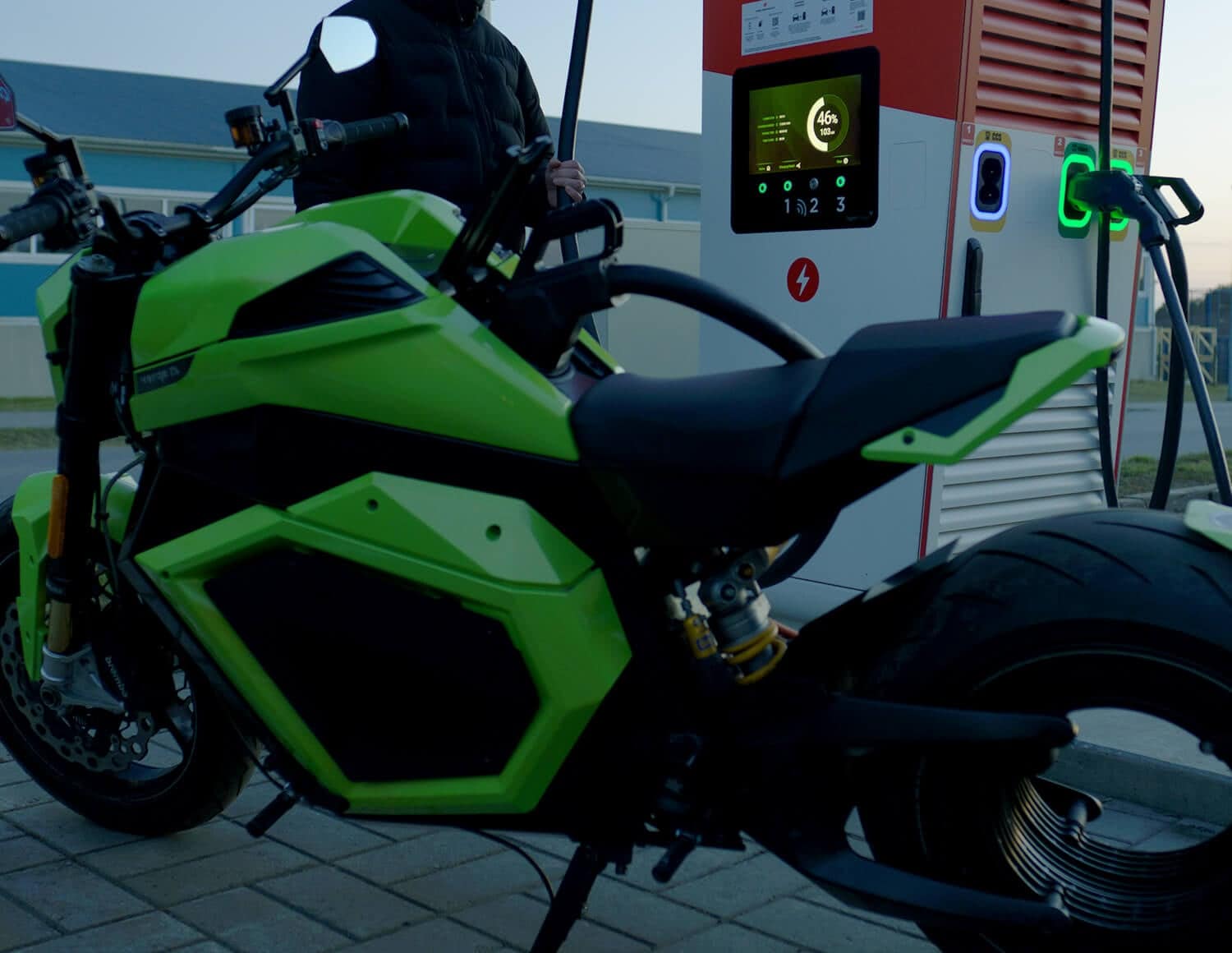 A bright green electric motorcycle is parked at a charging station while a person stands nearby. The display on the charger shows charging information. The scene takes place outdoors on a paved area.