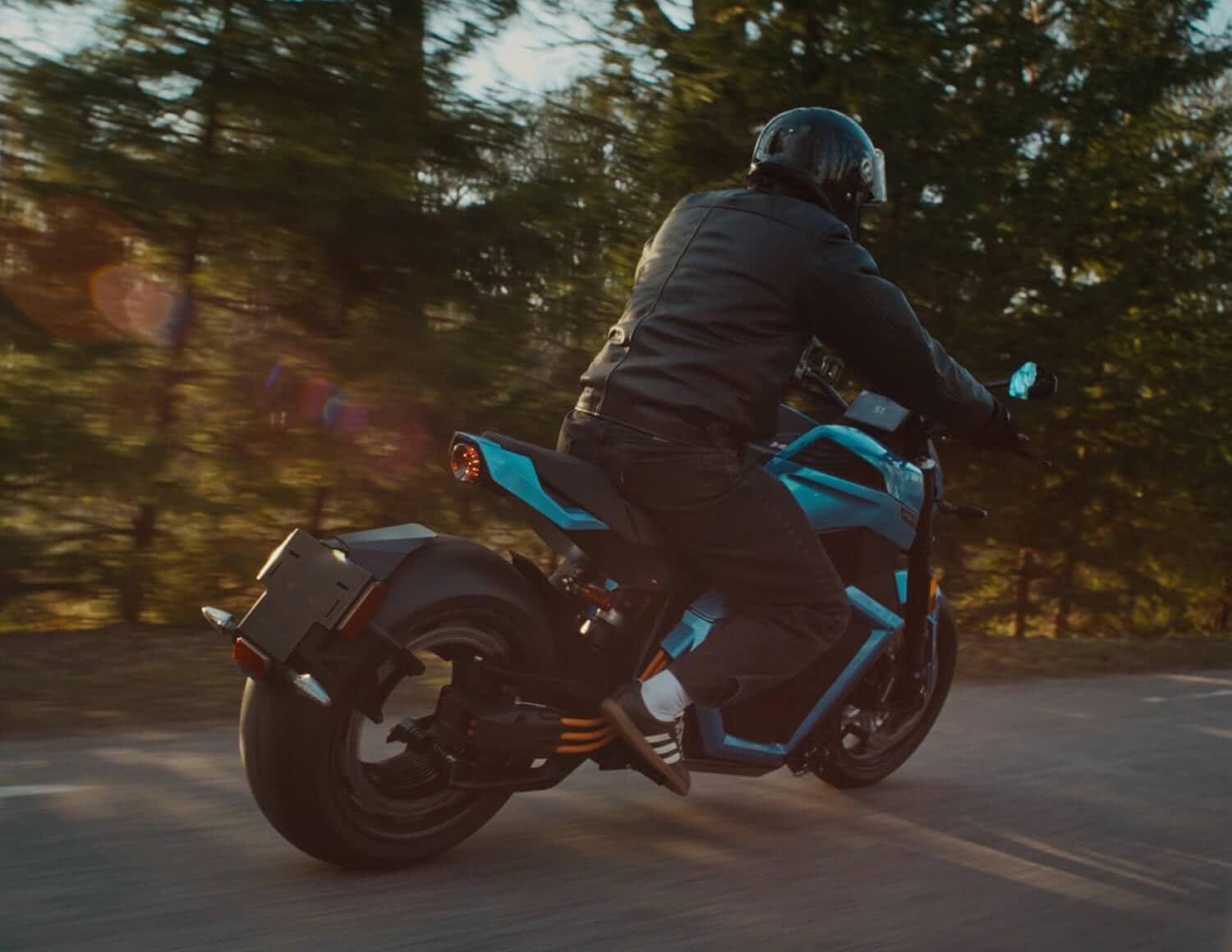 A person wearing a black helmet and jacket rides a blue electric motorcycle on a road surrounded by green trees, with sunlight filtering through the branches.