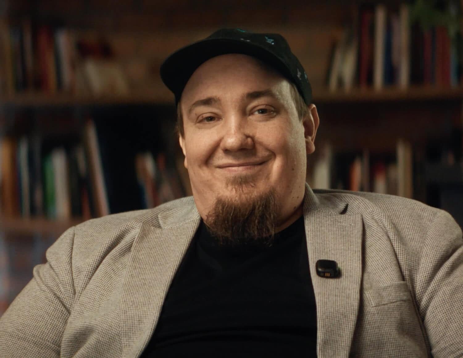 A man wearing a light gray blazer, black shirt, and black cap smiles at the camera. He has a short beard and is seated in front of a blurred background with bookshelves.