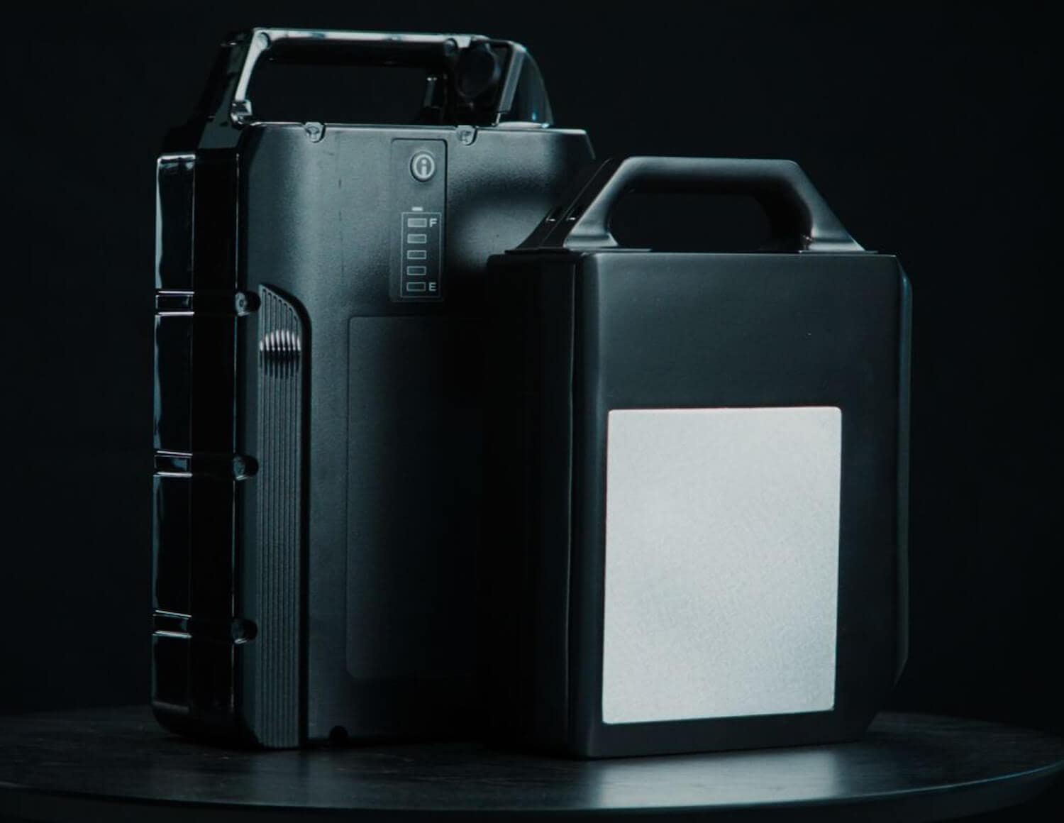 Two black, rectangular battery packs with handles are standing upright on a dark surface against a black background. One battery has a visible LED indicator and the other has a blank white label.