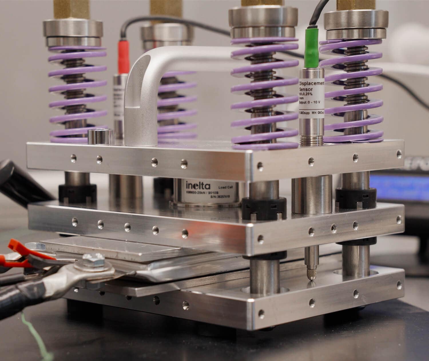 A close-up of a metal testing machine with purple springs, displacement sensors, and metal clamps holding a sample in place for compression or tension testing in a laboratory setting.
