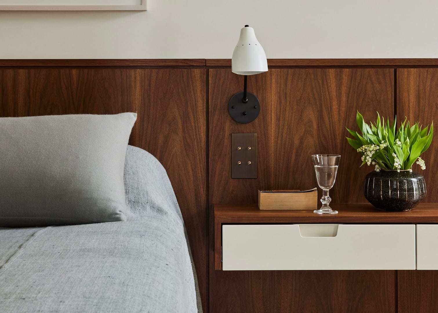 A neatly made bed with gray bedding is next to a wooden nightstand with a white drawer. The nightstand holds a vase with green plants, a glass of water, and a book. A white wall-mounted lamp is above the nightstand, against a wood-paneled wall.