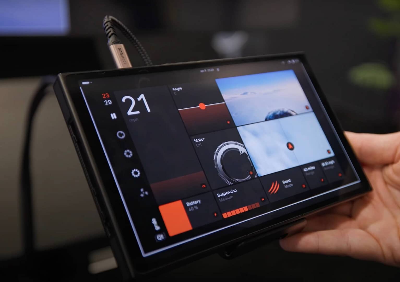 Close-up of a hand holding a black tablet device displaying a dashboard interface. The screen shows speed, angle, battery life, and other technical metrics with various dials and indicators against a dark background.