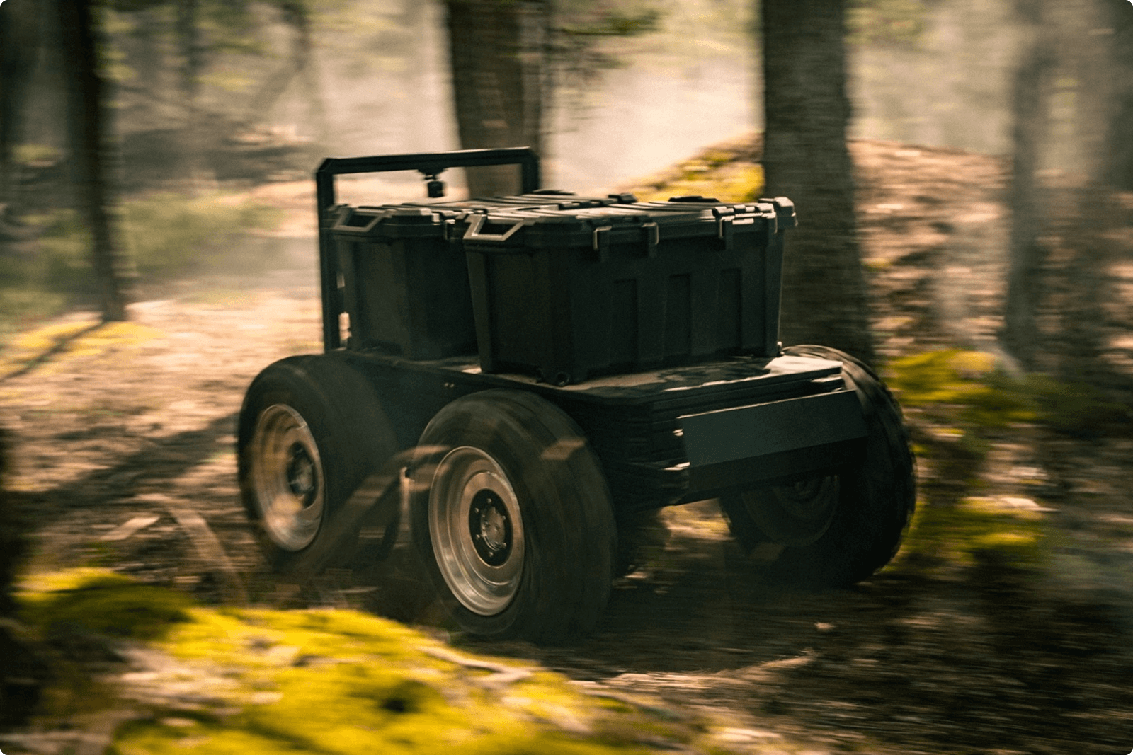 A rugged, four-wheeled robotic vehicle with a black storage container drives quickly through a misty forest, kicking up dirt and leaves as it navigates the terrain.