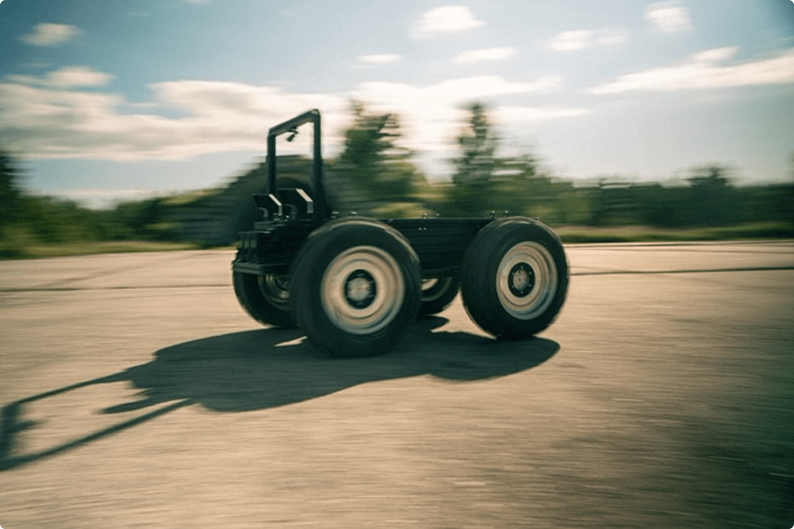 A four-wheeled robotic platform moves quickly across a paved surface outdoors, with blurred background indicating motion and trees visible in the distance.