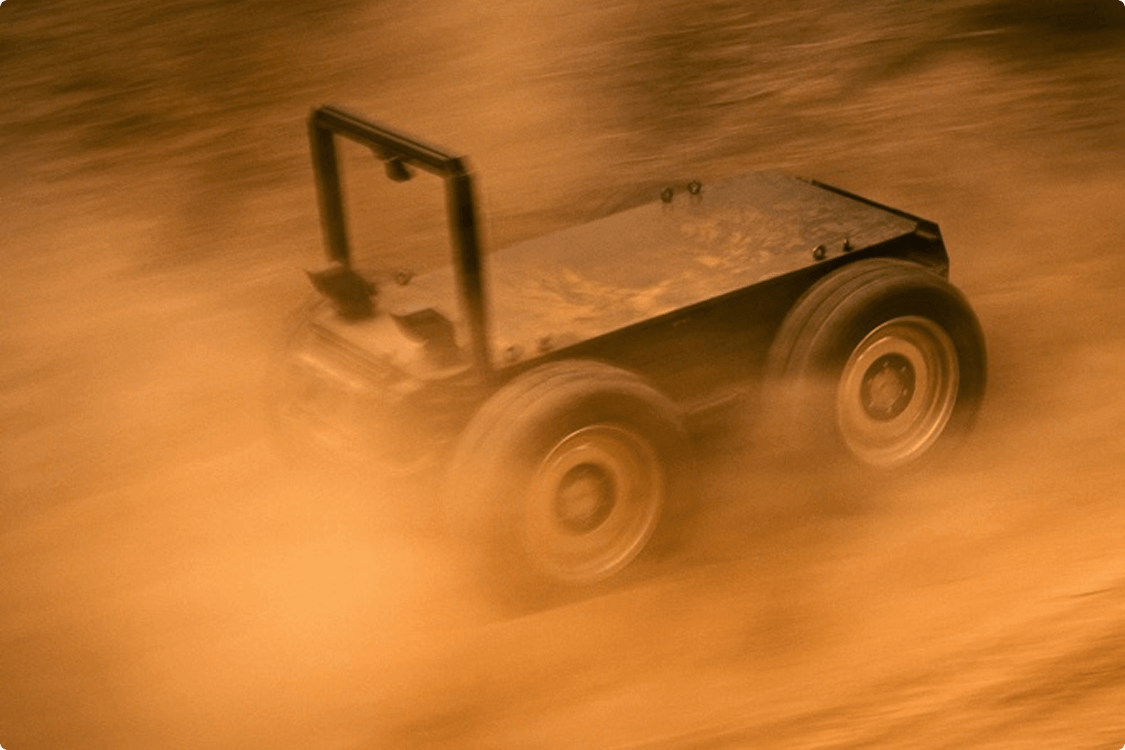 A small, rugged, four-wheeled robotic vehicle moves through a cloud of orange-brown dust, suggesting it is navigating rough terrain at speed.