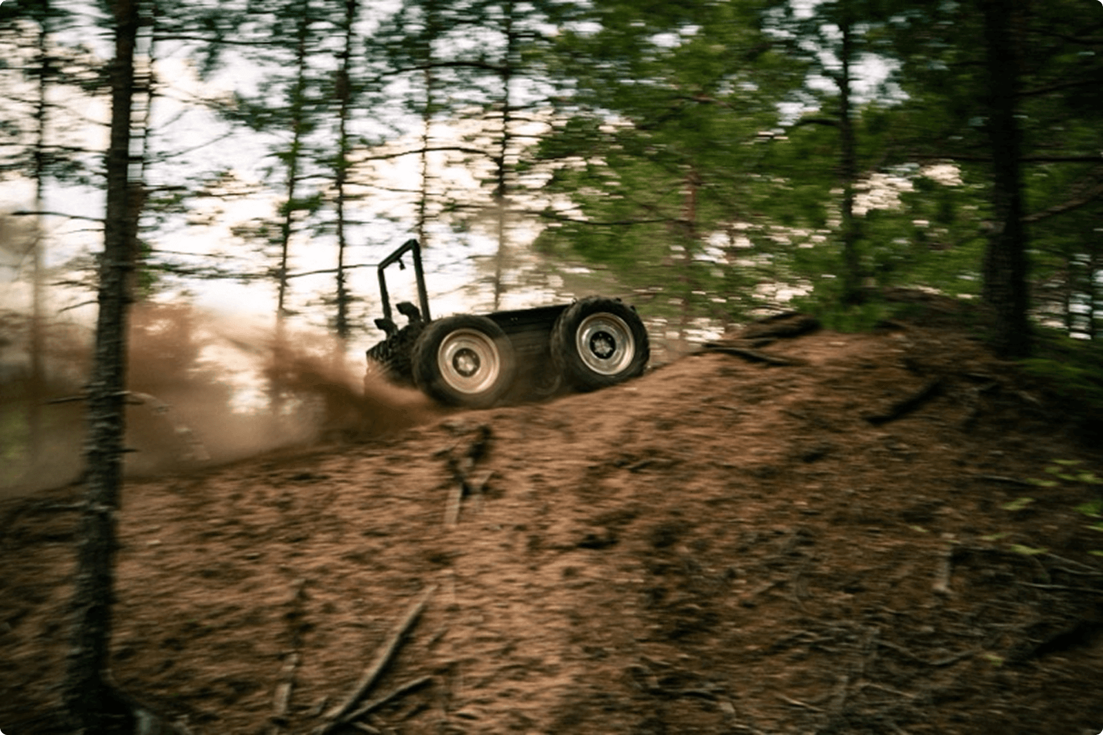 An off-road vehicle speeds up a steep dirt hill, kicking up dust, surrounded by tall trees in a forest setting.