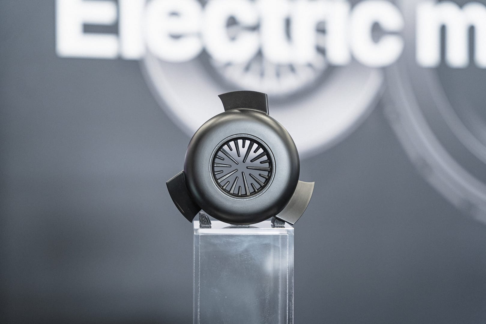 A black, circular device with three blades is displayed on a clear stand against a blurred background featuring the words "Electric m." The device has a central, intricate pattern and appears to be showcasing technology.