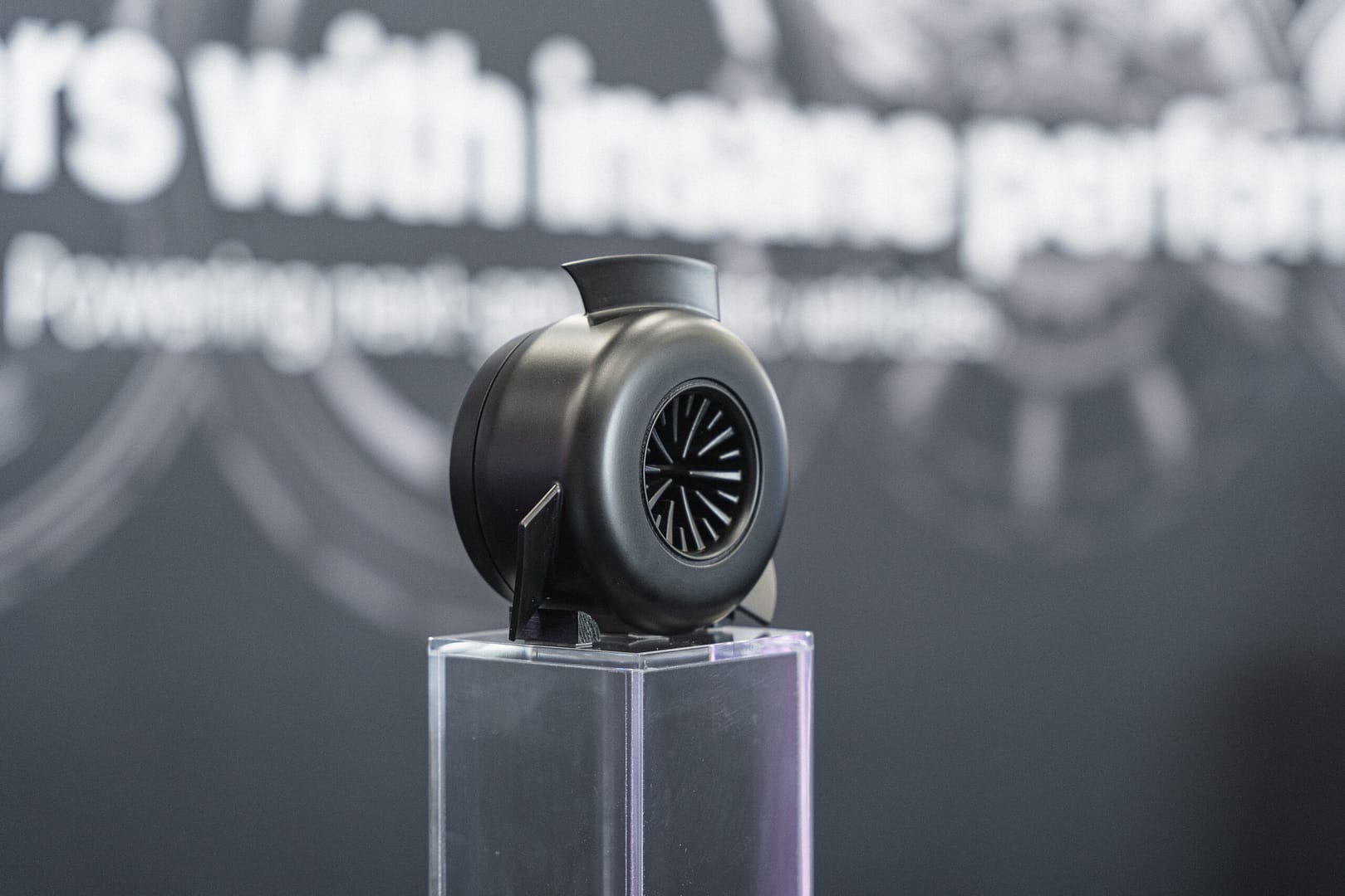 A black, turbine-shaped object displayed on a transparent stand against a blurred background with partial text. The object resembles an engine component, possibly a turbocharger.
