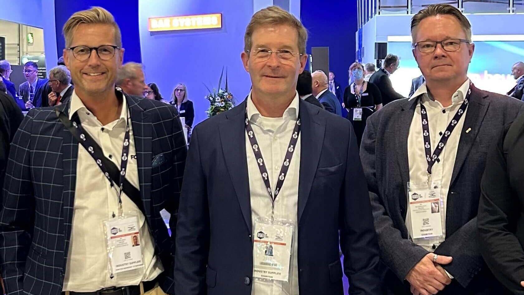 Fortaco-social-share-14.5.25-3 Three men wearing business attire and conference badges stand together, smiling, at an indoor event with other attendees and booths visible in the background.