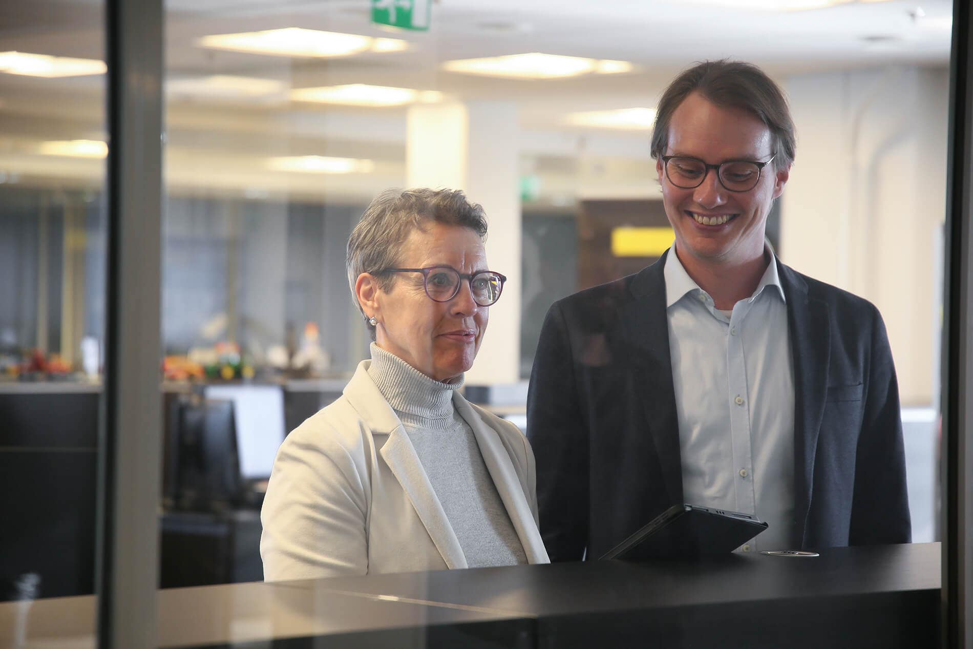Two people wearing glasses and business attire stand indoors, smiling and looking at a tablet together. They are seen through a glass partition in a modern office setting.
