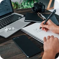 A person writes in a notebook beside a laptop, wireless earbuds, a smartphone, a camera, and a closed notebook on a desk.