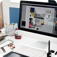 A desktop computer on a white desk displays a colorful graphic design. Nearby are pens, a notepad, eyeglasses, a graphics tablet, and a cup of coffee. The workspace appears organized and creative.