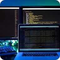 A workspace with a laptop and two monitors displaying lines of code, alongside a keyboard and mouse, illuminated by blue-green ambient lighting.