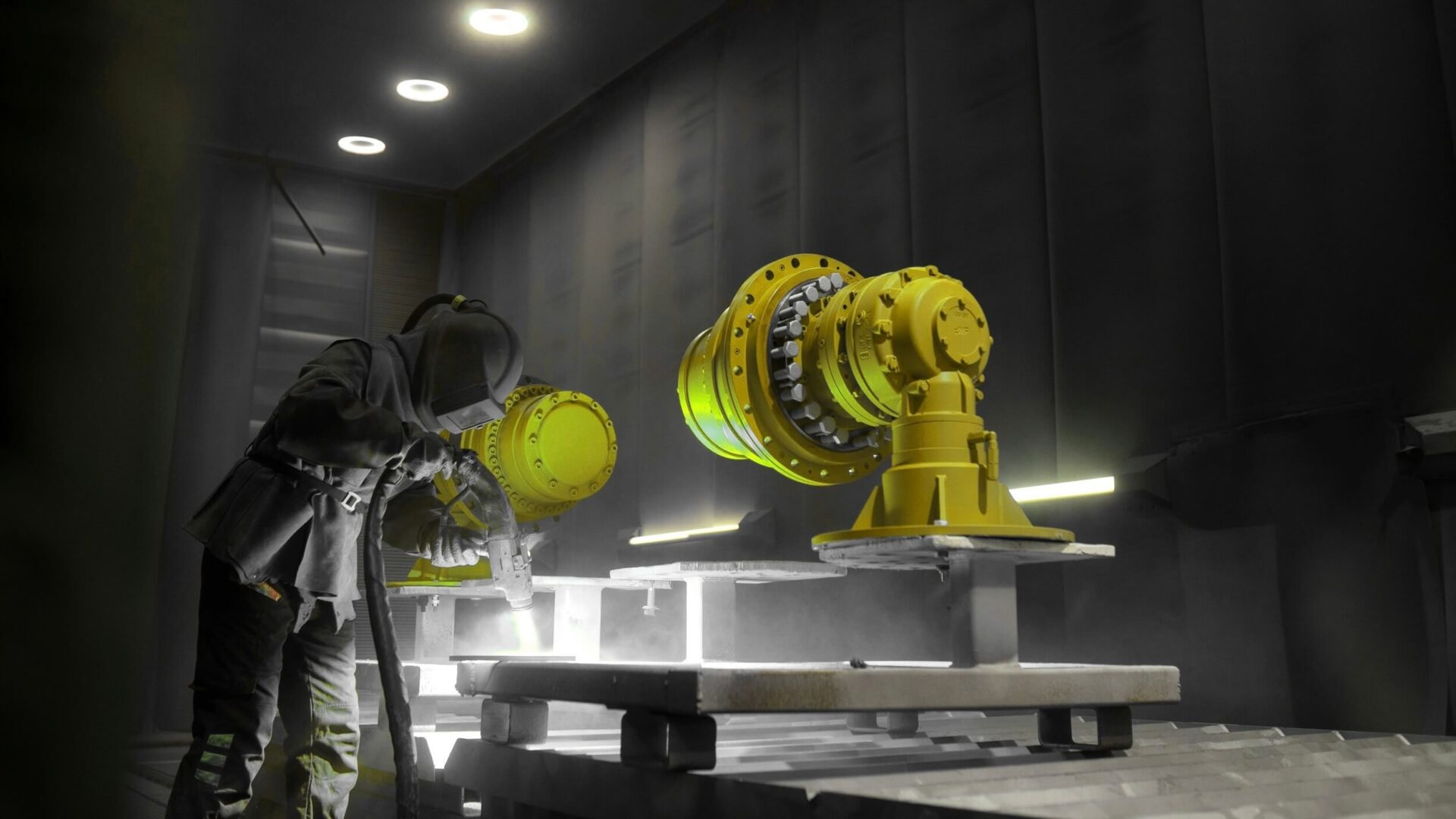 Fortaco-social-share-14.5.25-3 A worker in protective gear sandblasts or paints large yellow industrial machine parts inside a dimly lit workshop with bright overhead lights.