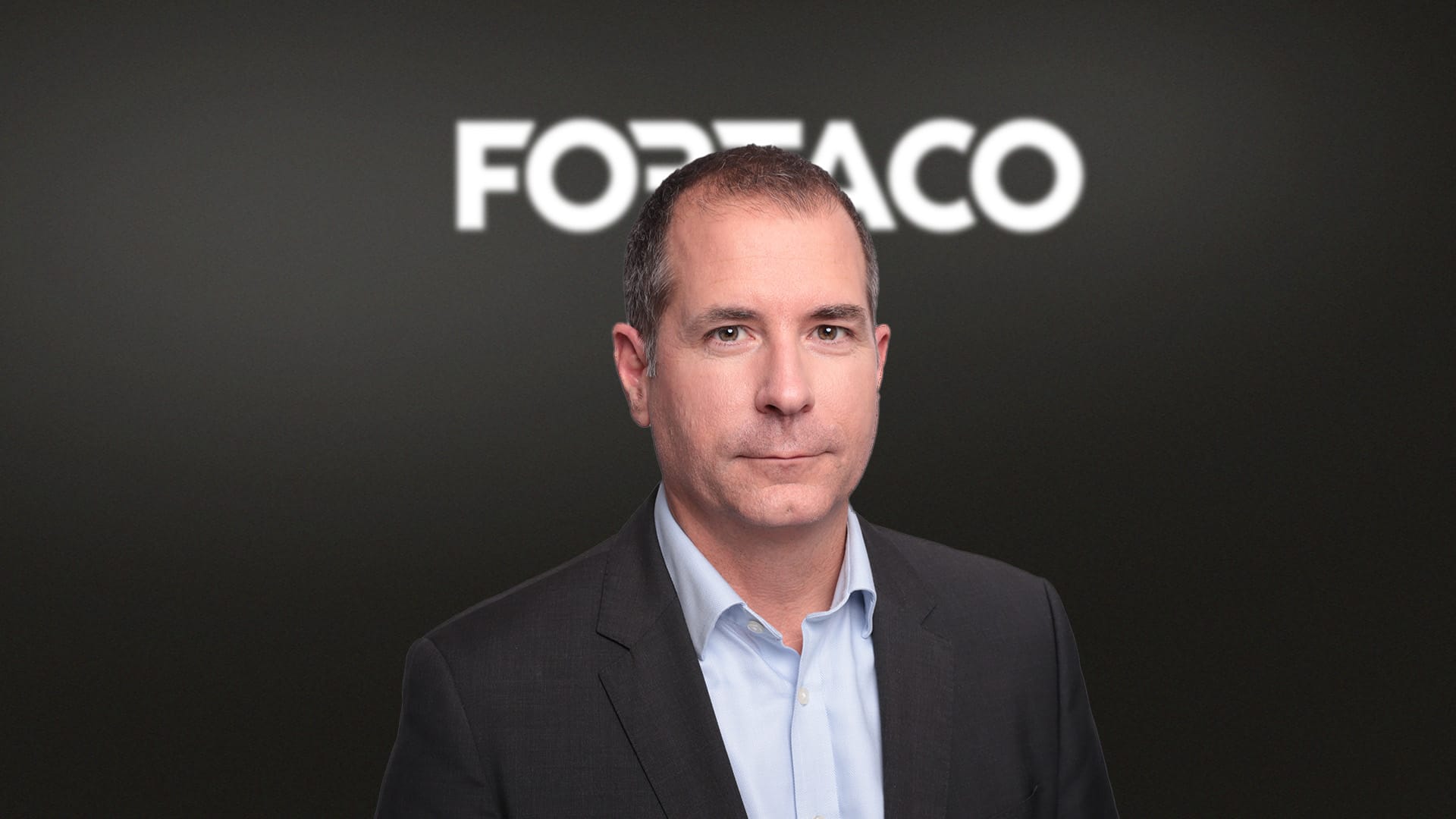 A man in a suit stands in front of a dark background with the word “FORTACO” in bold white letters behind him.