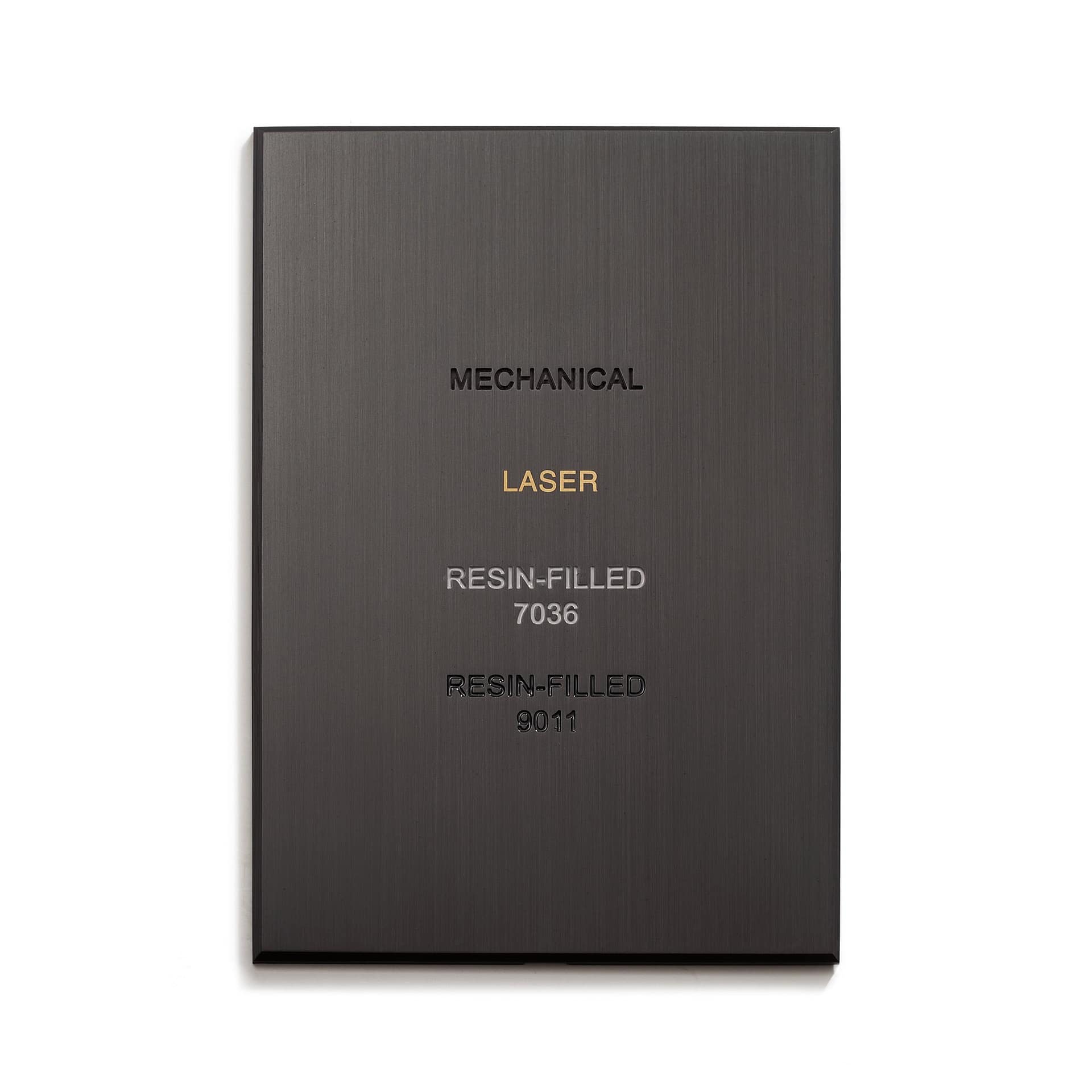 A close-up of a dark metallic plaque displaying different types of text inscriptions. The plaque has the words "MECHANICAL," "LASER," "RESIN-FILLED 7036," and "RESIN-FILLED 9011" engraved, showing different engraving techniques.