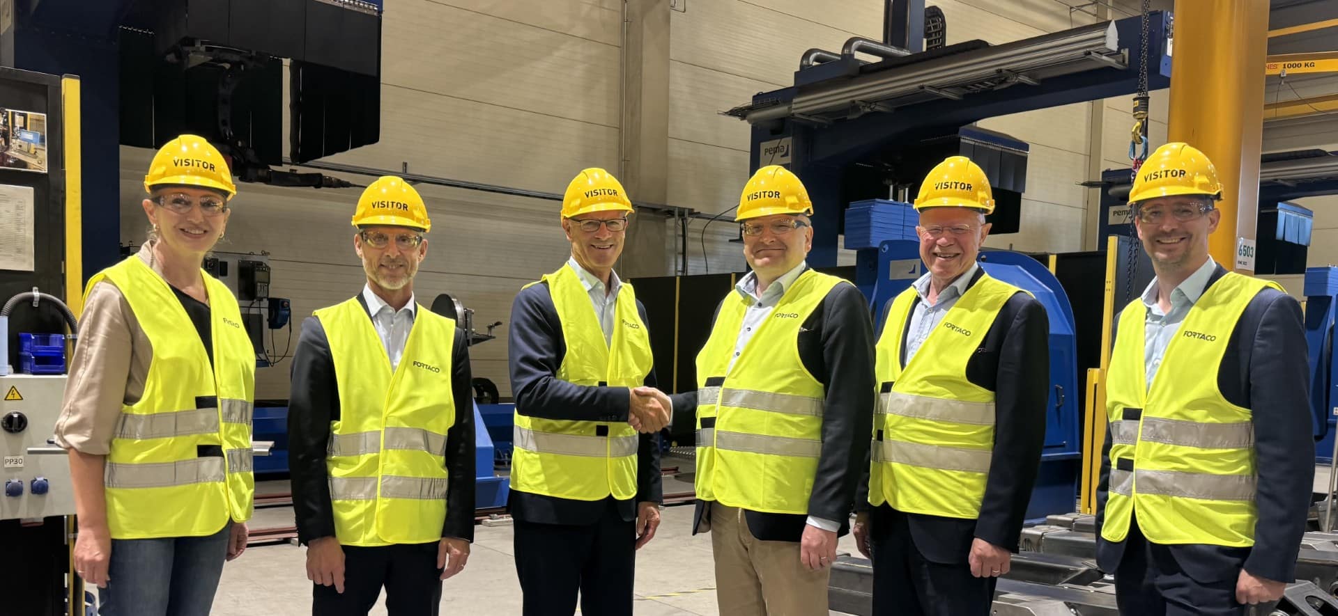 Six people in hard hats and yellow safety vests stand in a factory. Two men in the center are shaking hands, while others smile at the camera. Industrial equipment is visible in the background.