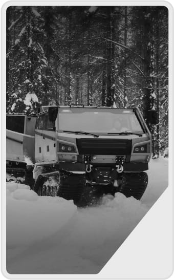 A tracked all-terrain vehicle drives through a snowy forest, surrounded by snow-covered trees and thick snow on the ground.