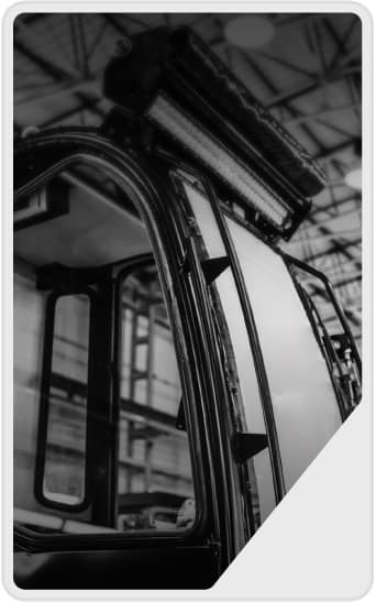 Black and white close-up of a vehicle's cabin window and roof, showing the frame and overhead lighting, with a blurred industrial ceiling in the background.