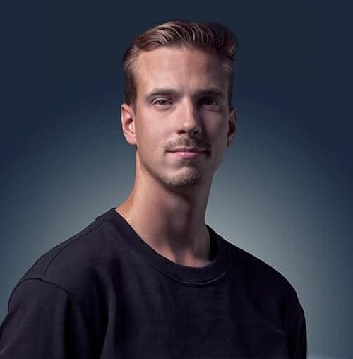 A young man with short, light brown hair and light facial hair wearing a black crew-neck shirt, posing against a gradient dark blue background.