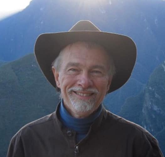 A man in a hat smiling in front of a mountain.