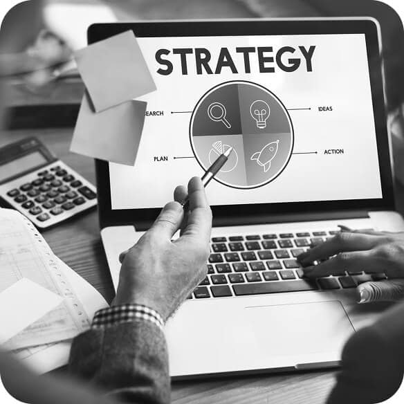 Black and white photo of two people discussing a strategy diagram on a laptop screen with four sections: research, ideas, plan, and action. A calculator, papers, and sticky notes are on the desk.