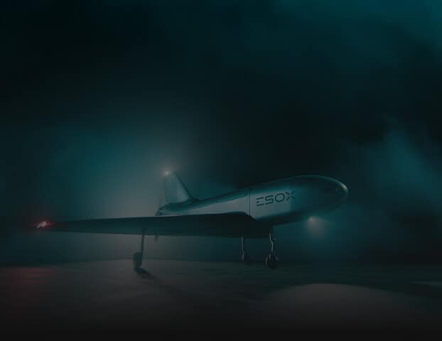 A sleek, unmanned aircraft labeled "ESOX" is illuminated by soft blue lighting in a dark, foggy environment, with its landing gear deployed and navigation lights glowing.