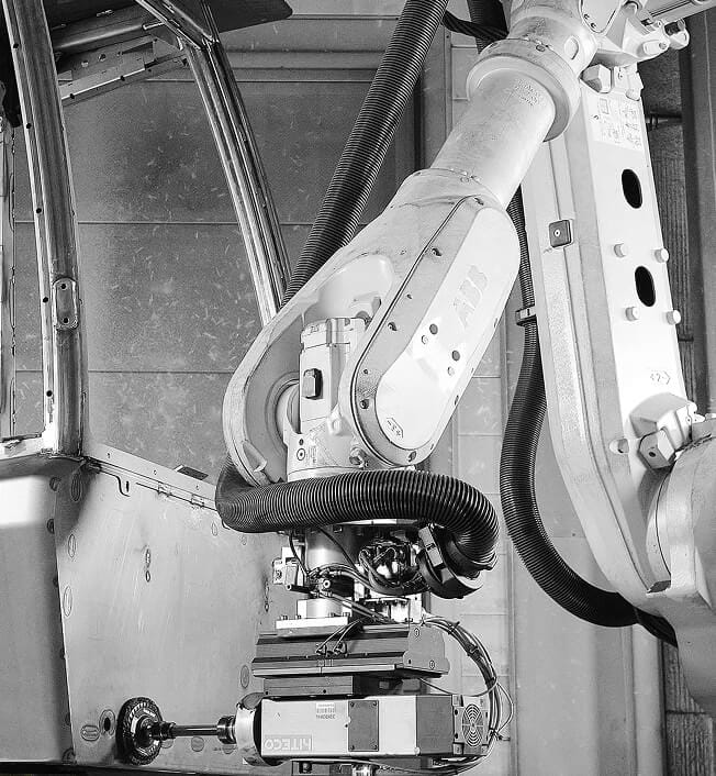 A large industrial robotic arm equipped with various attachments is working inside a factory setting, handling machinery components. The environment appears clean and metallic.