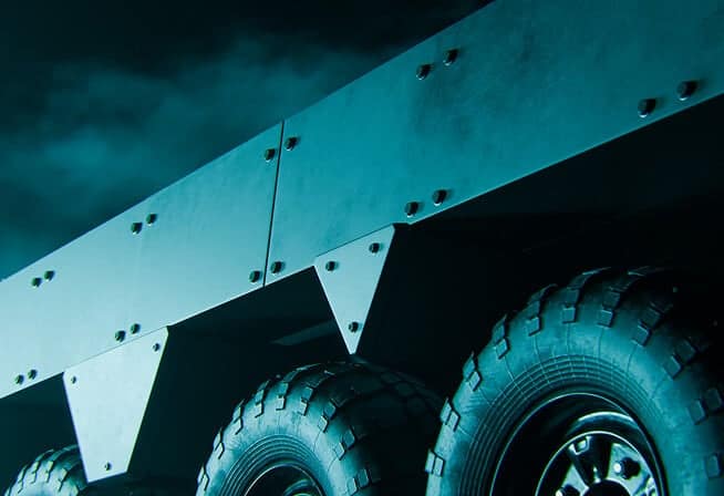 Close-up view of large, rugged off-road vehicle tires and metallic armored panels, illuminated by teal lighting with a cloudy, dark sky in the background.