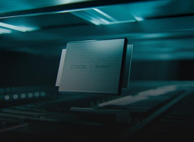 A sleek, metallic battery pack labeled "ESOX Battery" is displayed in a futuristic, teal-lit environment, positioned above parallel rails or conveyor belts.