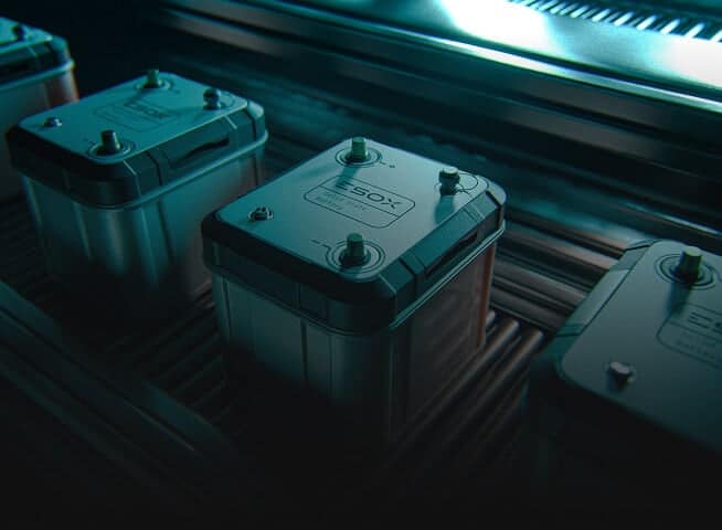 Three rectangular batteries with terminals on top sit on a conveyor belt under blue lighting in an industrial setting. The battery in the center has a label reading “ESOX.”.