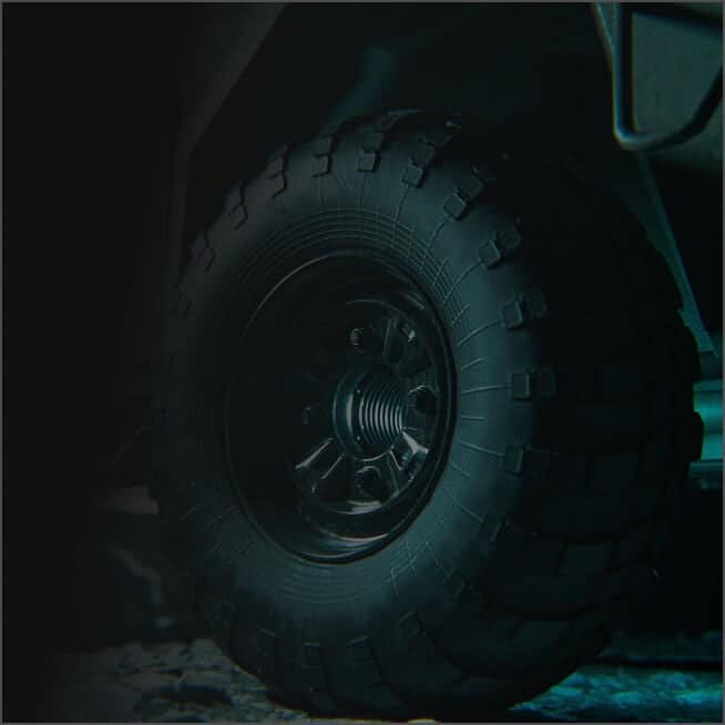 Close-up of a rugged, black off-road vehicle tire with deep tread patterns, mounted on a dark metal wheel, on a rough surface with dim lighting.
