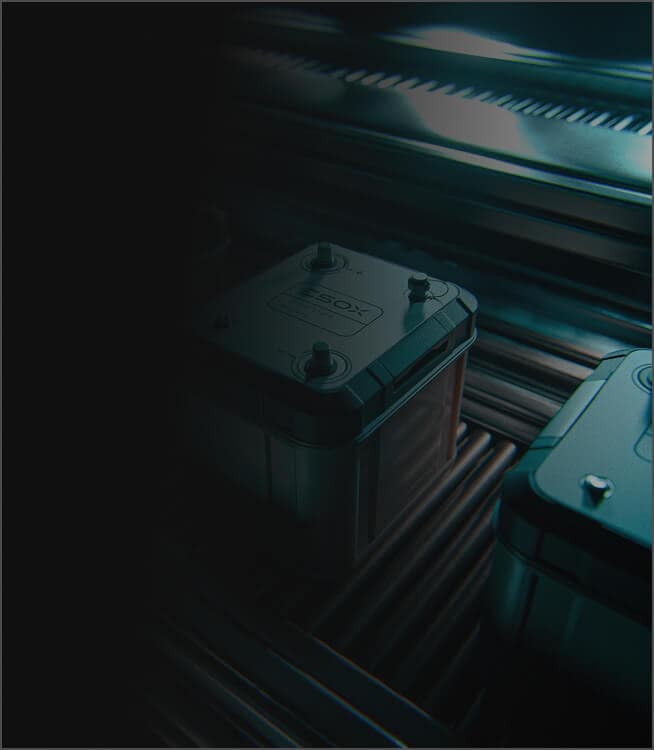 A dark, moody photo showing two industrial batteries or power cells on a metallic, ribbed surface under dramatic blue lighting. The left side of the image is mostly in shadow.