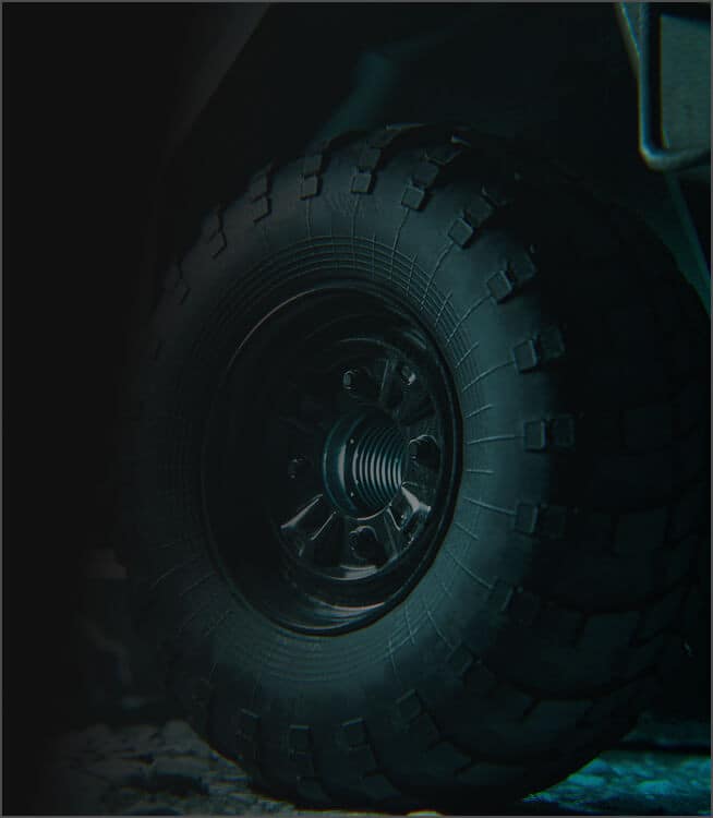 Close-up view of a large, rugged off-road vehicle tire with deep treads, partially illuminated against a dark background. The focus is on the texture and pattern of the tire.