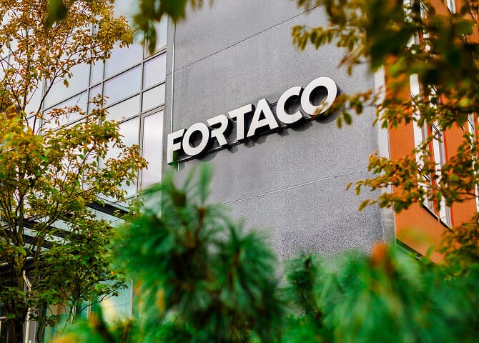 The image shows the exterior of a modern building with the word "FORTACO" displayed in large white letters on a grey wall, partially framed by green foliage and trees.