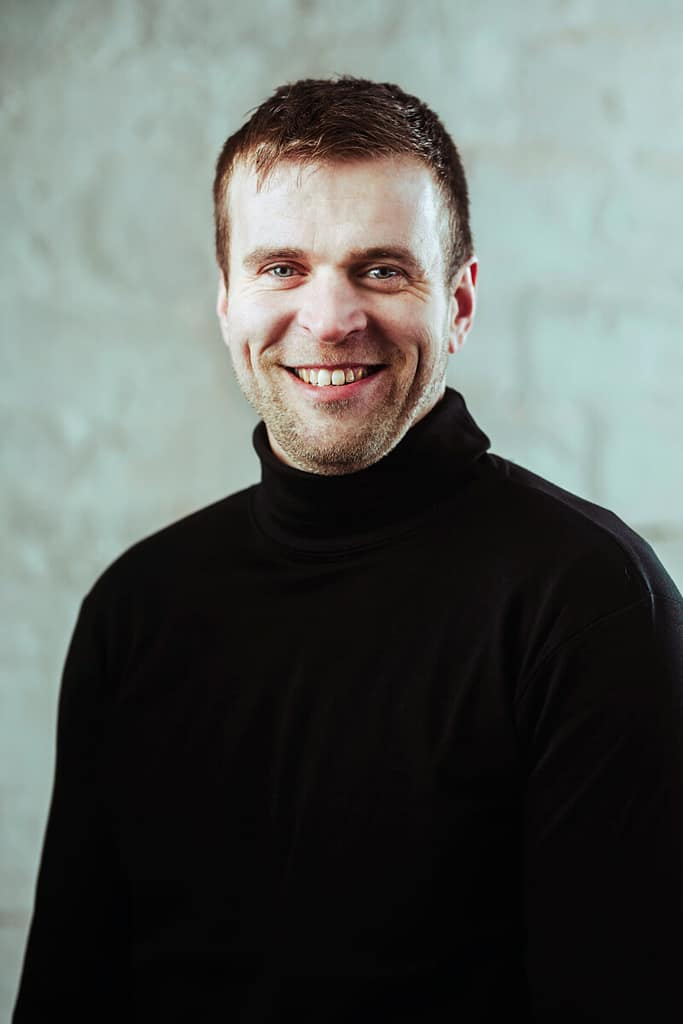 Mies, jolla on lyhyet ruskeat hiukset ja parta, hymyilee kameralle. Hänellä on yllään musta villapaita, jonka taustalla on kuvioitu harmaa seinä.