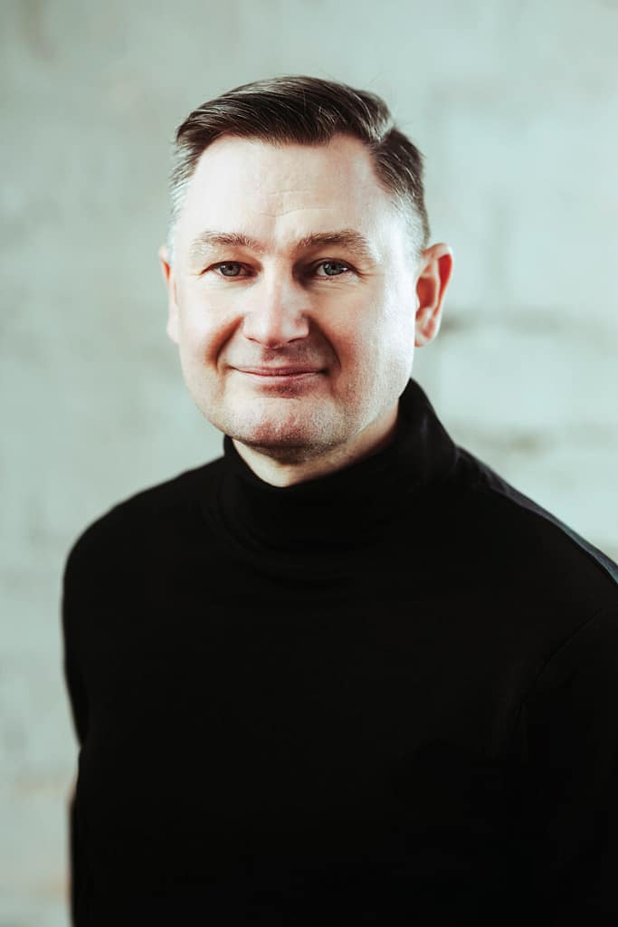 Keski-ikäinen mies, jolla on lyhyet, siististi muotoillut hiukset ja jolla on musta villapaita, poseeraa vaaleaa taustaa vasten. Hän hymyilee hieman ja katsoo suoraan kameraan.