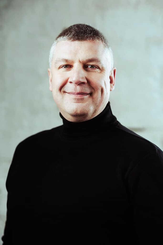 En man med kort grått hår ler medan han bär en svart polokrage mot en strukturerad grå bakgrund.