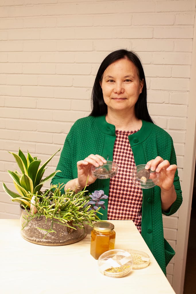 A woman in a green cardigan and plaid shirt sitting at a table, smiling at the camera while holding two small jars of seeds with a potted plant arrangement beside her.