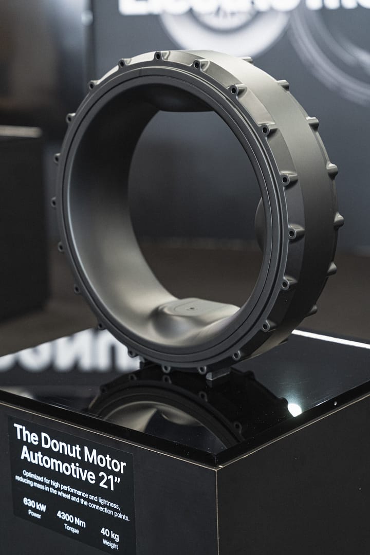 A large, circular automotive component called "The Donut Motor Automotive 21" is displayed. It is mounted on a black platform, showcasing specifications: 230 kW, 4300 Nm, 40 kg. The background is blurred.