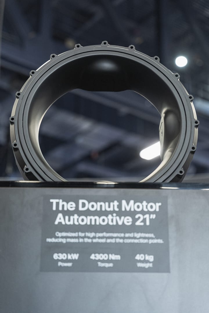 Close-up of a large, circular automotive component named "The Donut Motor Automotive 21" on display. The sign below reads: "630 kW Power, 4300 Nm Torque, 40 kg Weight." The background is blurred with overhead lights.