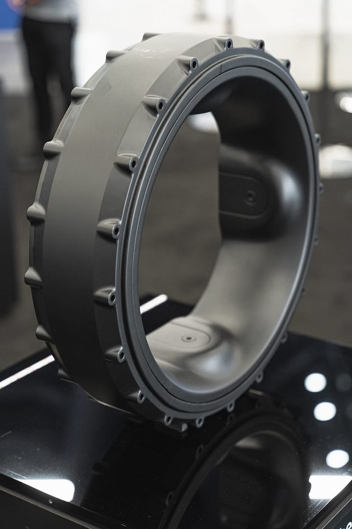 A large, circular industrial component with a smooth inner surface and evenly spaced protrusions on the outer edge. It is displayed on a glossy black platform in a brightly lit setting.