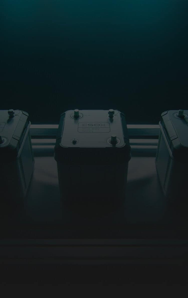 A dark, moody image of a battery pack with two metal terminals on top, placed on a metallic surface, with two similar objects partially visible on either side.