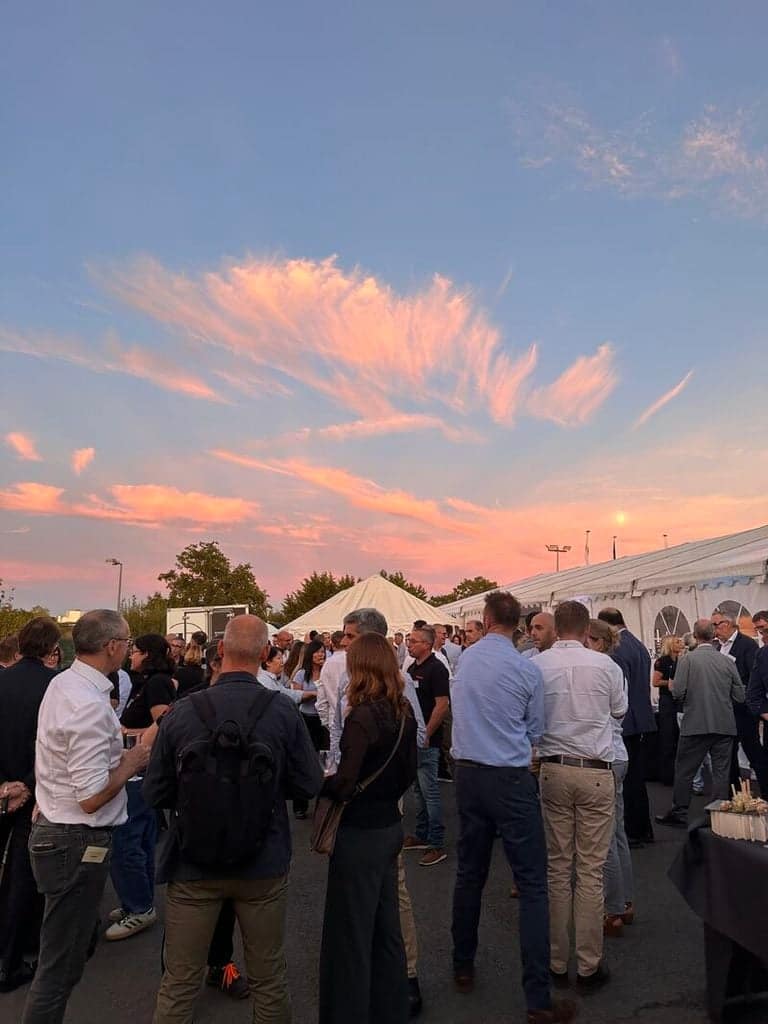 A group of people socializing at an outdoor event under a pink and orange sunset.